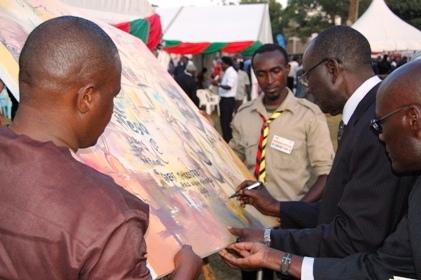 Mr. Mutekanga Igaga (R) assisted by his son Mr. Daudi Mutekanga (R) and Mr. Agaba Issa Mugabo (L) signs the Mak@90 Grand Finale commemorative live painting on 3rd August 2013 at the Freedom Square, Makerere University, Kampala Uganda. 3rd Right is the artist Rolands Tibirusya.